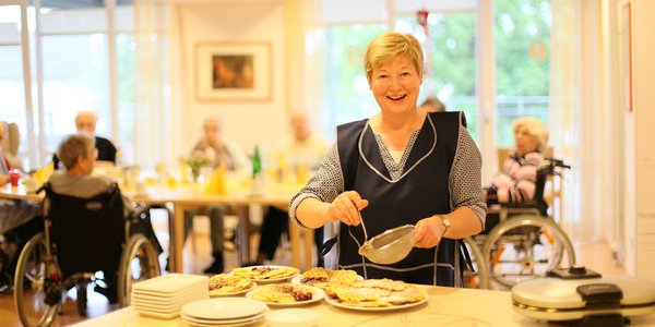 Eine Frau backt Waffeln, im Hintergrund ist die Kaffeetafel gedeckt.